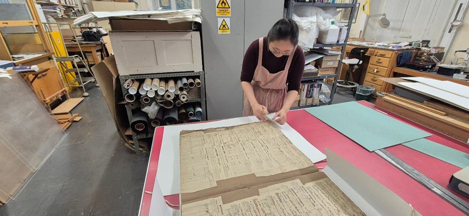 Person ymwneud â gwaith atgyweirio dogfen hanesyddol yn archif, yn rhoi sylw i'r i'r tudalennau'r hen gofrestr.