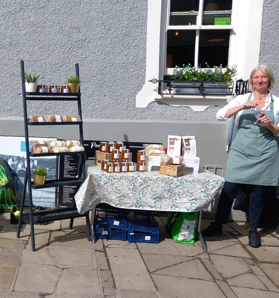 A vendor at the Conwy Honey Fair stands at a market stall filled with jars of honey, baked goods, and other products, with a decorative tablecloth and potted plants, in front of a grey building.