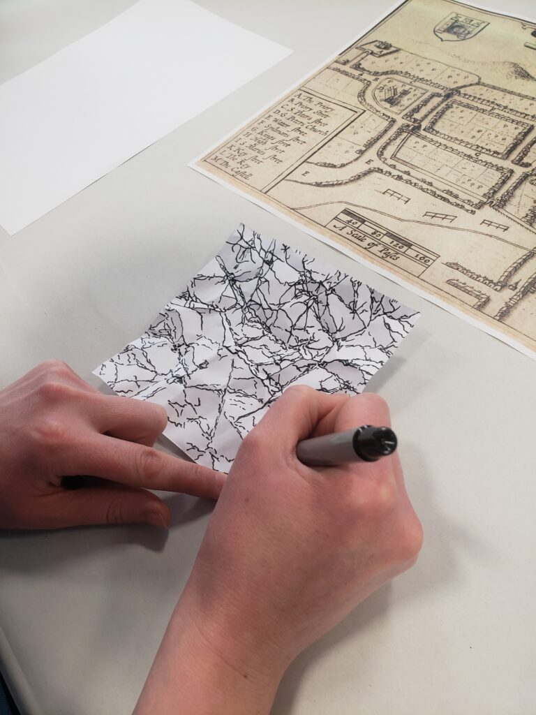 Mae dwylo'n llunio map mewn pen ar bapur gwyn, gerllaw mae copi o gynllun archifol.