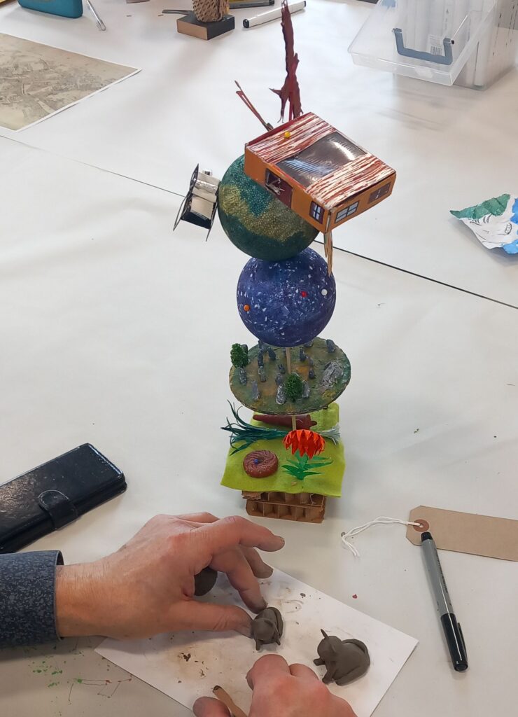 Hands are shaping small animals in clay. Nearby is a tall, stacked model featuring spheres, and various miniature landscapes.